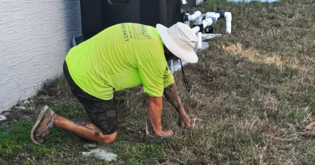 Brandon-Connolly-laying-sod | Connolly Landscaping Shane Connolly kneels with both hands in a patchy lawn beside a house, an outdoor unit and white pipes.
