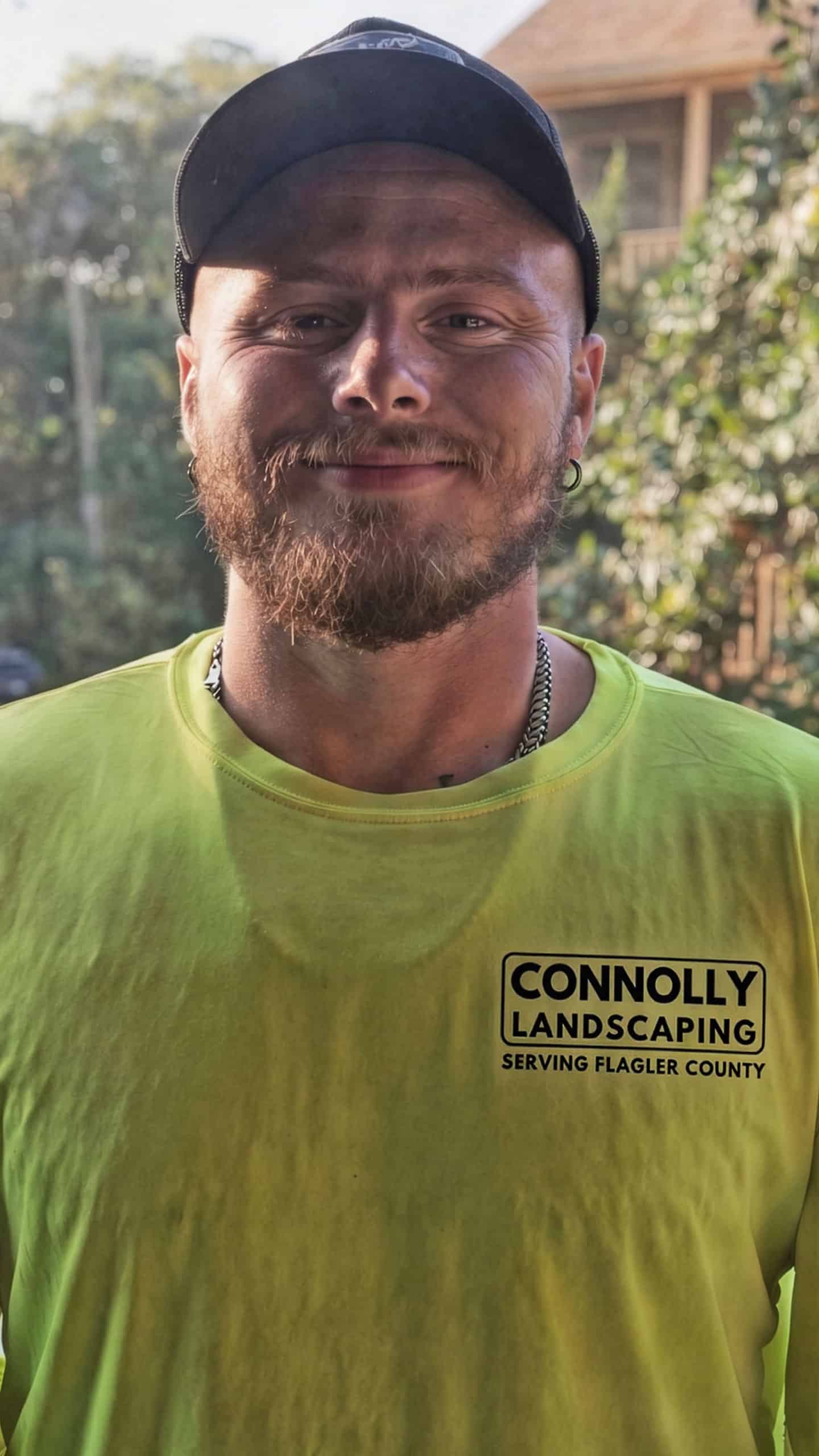 A man with a beard and cap smiles at the camera, wearing a bright yellow shirt that says Connolly Landscaping Serving Flagler County. Trees and a wooden building are visible in the background.