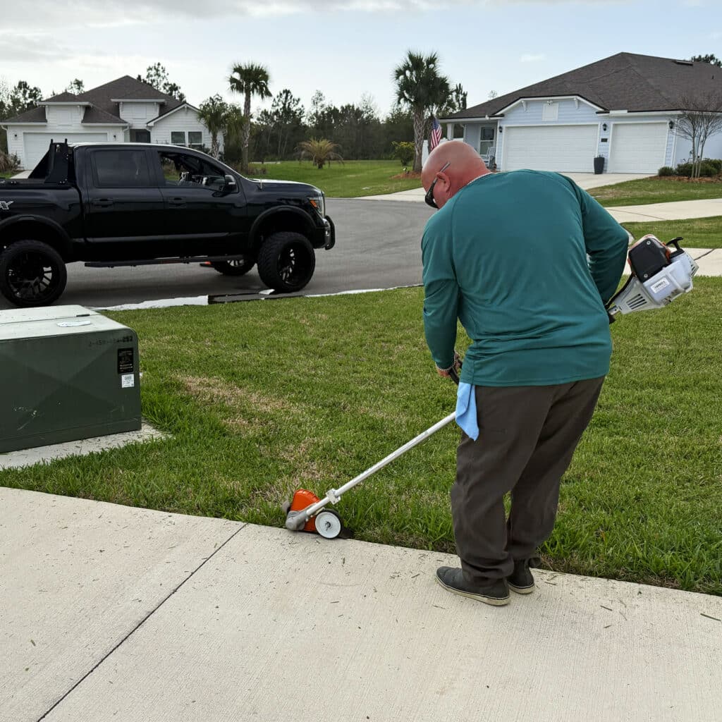 Keith-Connolly-Edging-Palm-Coast-Lawn | Connolly Landscaping A person wearing a green shirt uses a grass edger on the edge of a lawn next to a sidewalk in a suburban neighborhood. A black pickup truck and single-story houses are visible in the background.