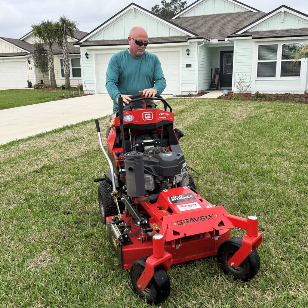 Keith-Connolly-Mowing-Palm-Coast-Lawn | Connolly Landscaping A man wearing sunglasses and a teal long-sleeve shirt mows a front lawn with a red Gravely walk-behind mower in front of a light blue house with a driveway and palm trees.