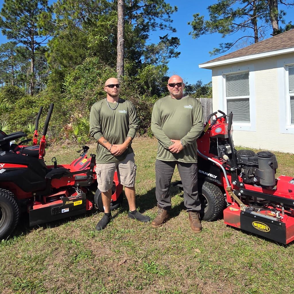 Two men in matching green long-sleeve shirts and sunglasses stand on grass between two red lawnmowers, with trees and a white building in the background on a sunny day.