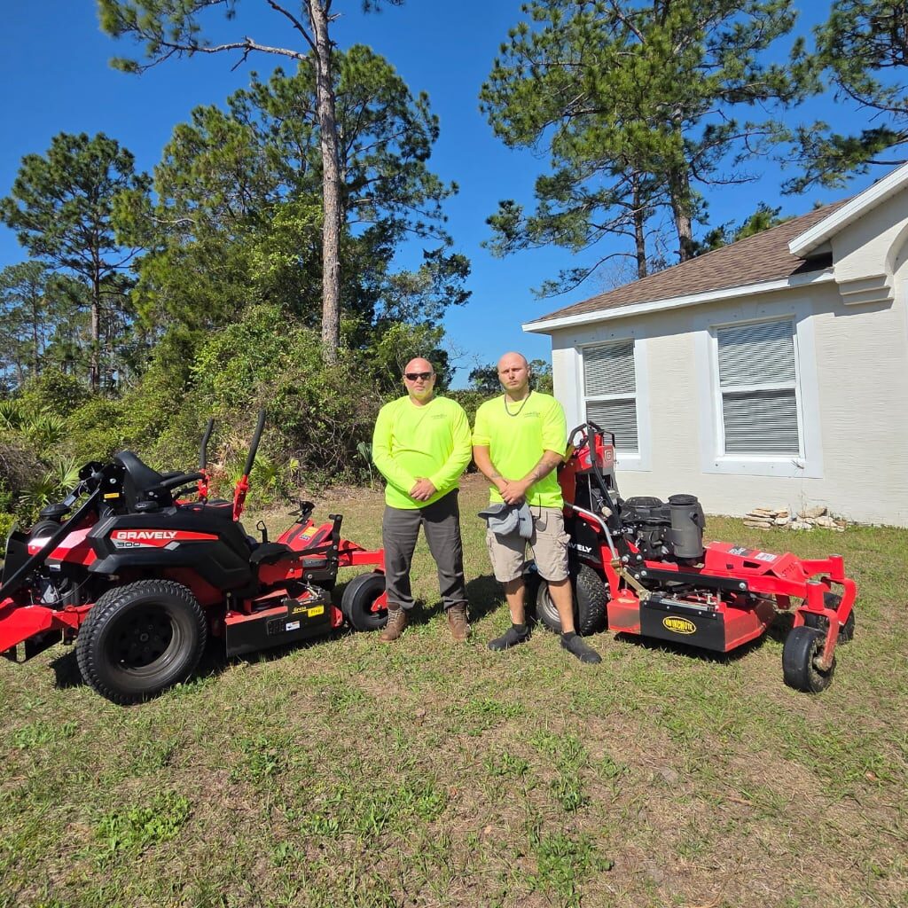 Two men in neon yellow shirts and shorts stand side by side on grass next to a red ride-on mower and a red walk-behind mower, with trees and a white house in the background under a clear blue sky.