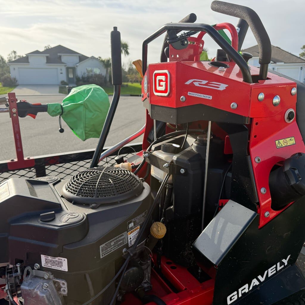 A close-up of a red Gravely RZ lawn mower parked on a residential street, showing details of the engine and controls, with houses and trees visible in the background.