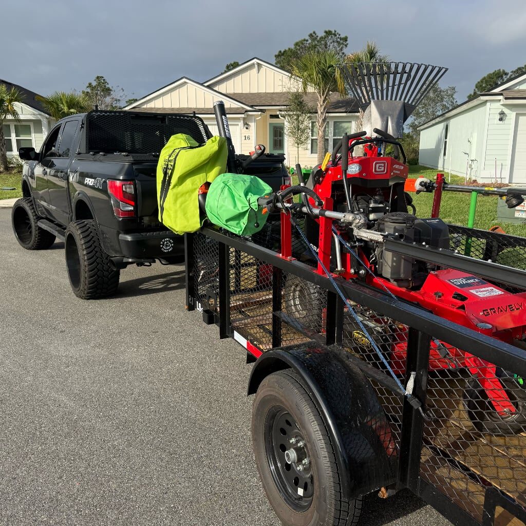 A black pickup truck with a trailer attached is parked on a street in front of houses. The trailer carries lawn care equipment, including a red mower, leaf blower, large rakes, and green bags.