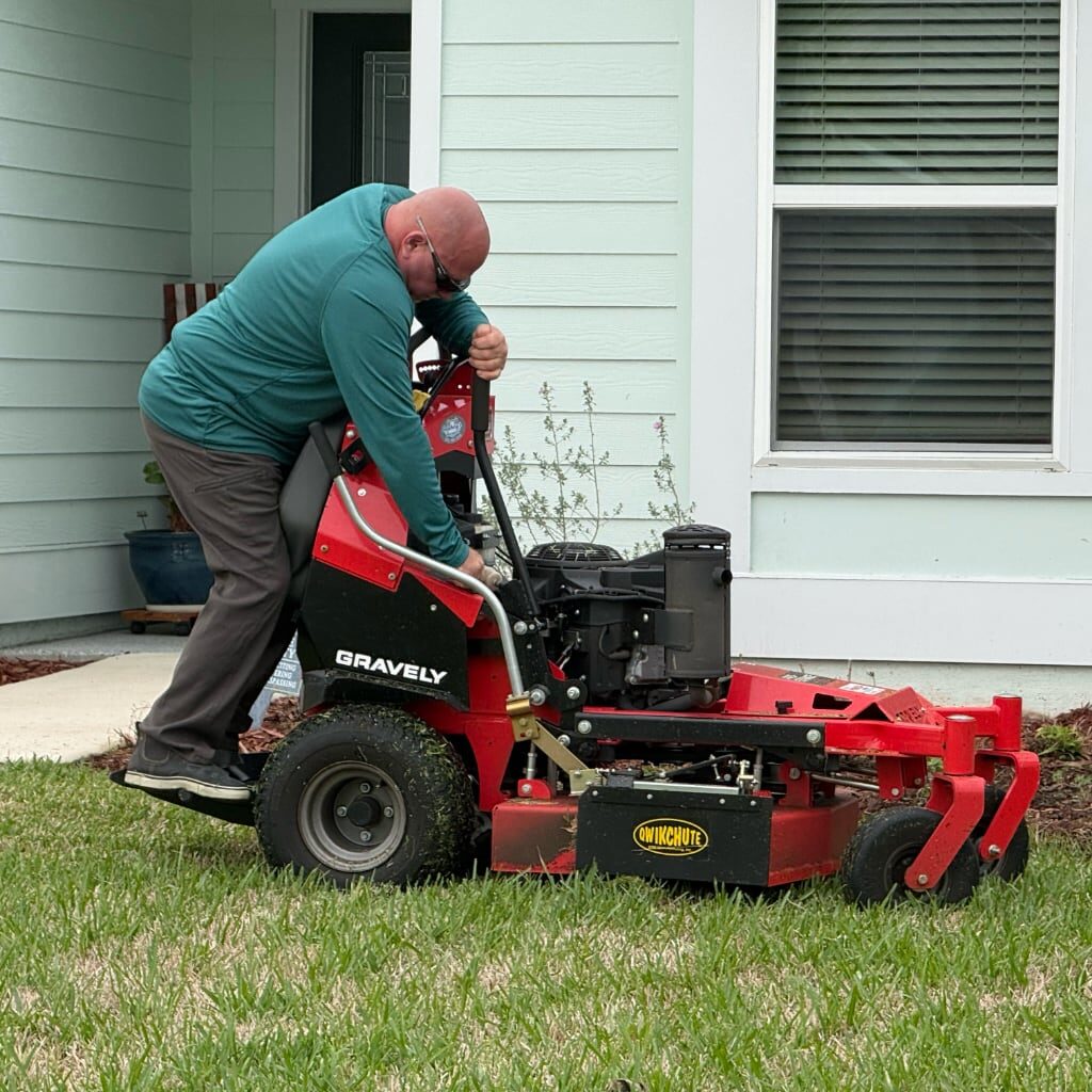 A man in a teal shirt and gray pants is starting a red Gravely lawn mower on a grassy lawn in front of a light-colored house with white trim and large windows.