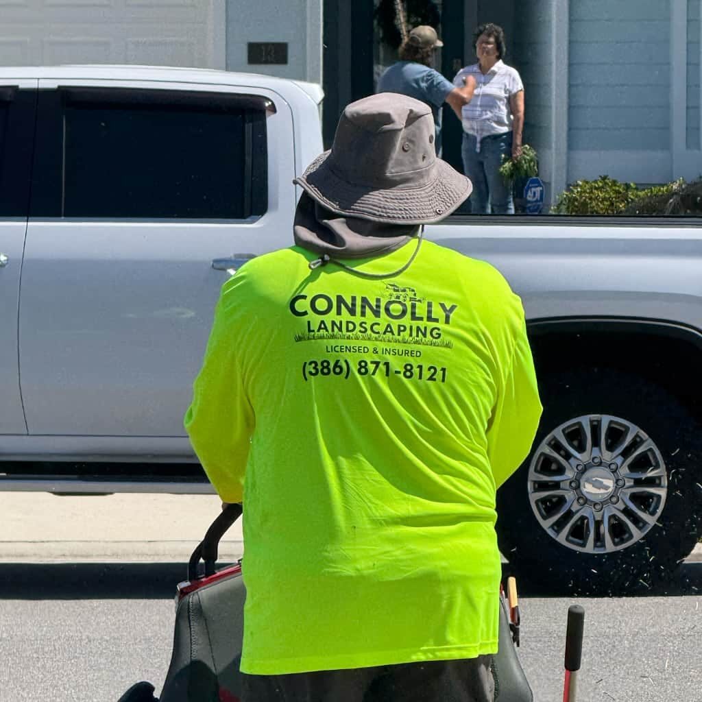 A person wearing a bright yellow Connolly Landscaping shirt mows a lawn near a street. In the background, a pickup truck is parked and two people are talking in front of a house.