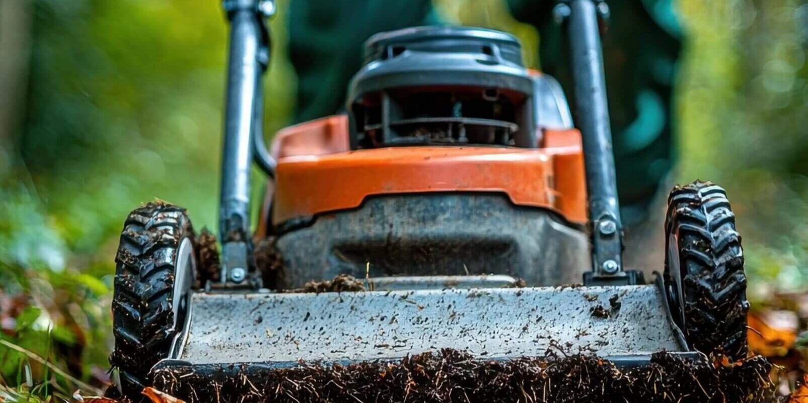 A person in green work clothes and white gloves operates an orange lawn mower, cutting through grass and leaves in an outdoor, wooded area. The photo is taken from ground level, emphasizing the mower’s front.
